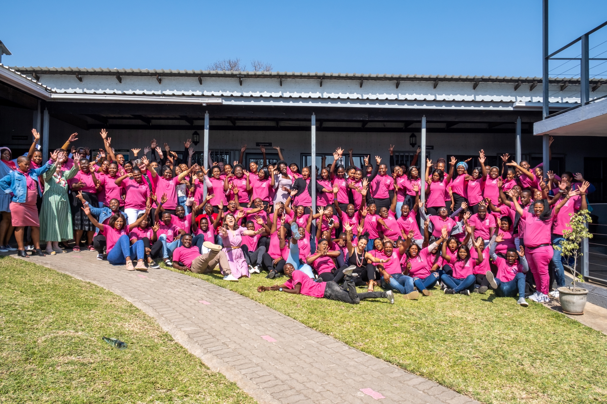 Felix-Studios-GWF-Aug-2022-144 A massive group of GWF team members and students, dressed in bright pink, gather outside a building, raising their hands and cheering in celebration under the clear blue sky.