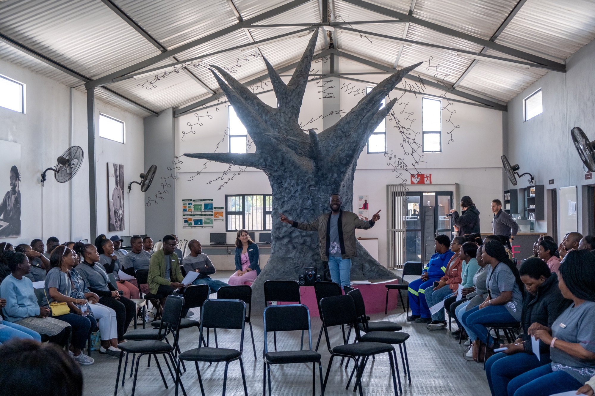 Felix-Studios-GWF-Aug-2022-161 A speaker stands confidently in front of a group of engaged listeners, gesturing animatedly, with a striking tree sculpture behind him as a symbol of growth and learning.