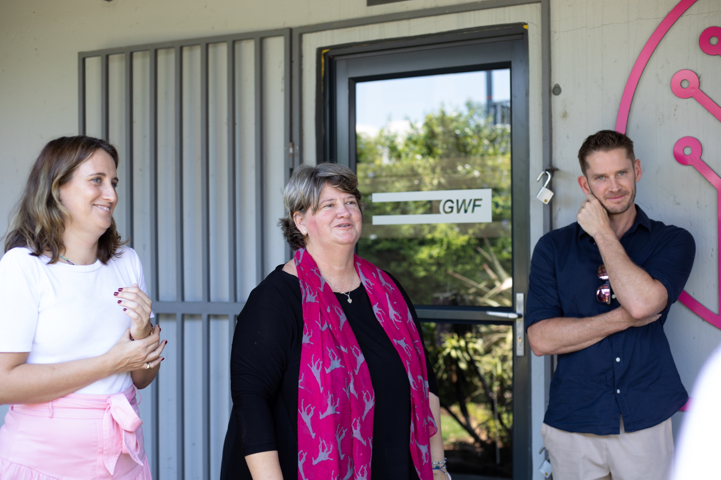 Marlin-Clark-rhino-africa-marketing-team-visit-at-gwf-9 Three individuals stand outside a GWF building, listening attentively, with one woman in a black outfit and a pink scarf speaking passionately while the others react with smiles and thoughtfulness.