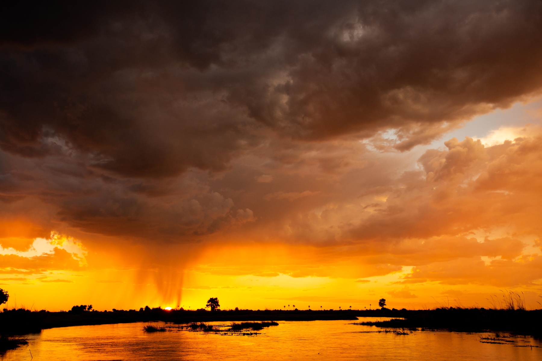 Duba-Plains-Camp-okavango-sunset-and-storm Dark storm clouds roll over the Okavango Delta as the setting sun paints the sky in fiery hues of orange and gold, with streaks of rain falling in the distance.